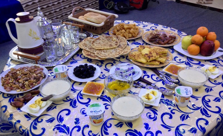 Large round table with various plates and dishes, containing pastries, bread, fruit, jams, cheese, semolina porridge, yoghurt. Also tea and coffee.