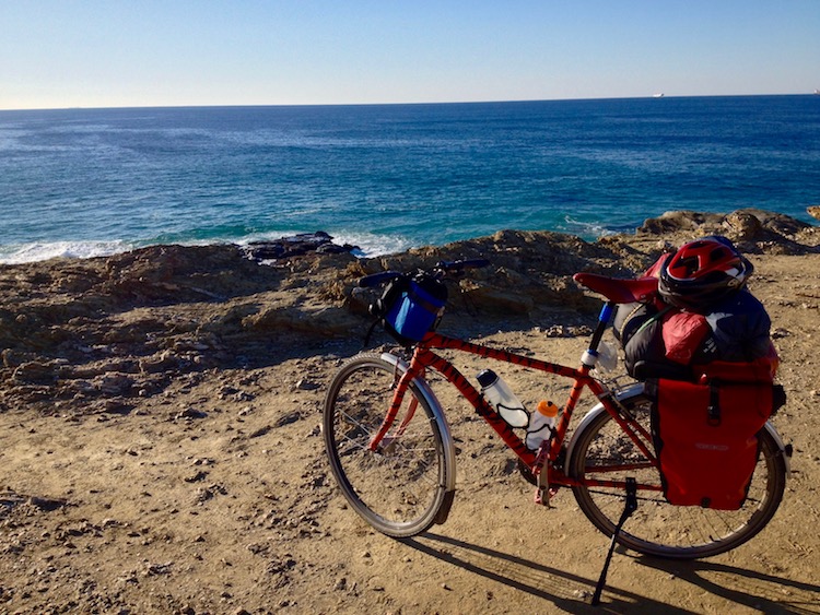 Dans bike, propped up on the edge of a golden, sandy cliff, overlooking deep blue waters of the Atlantic, with blue skies overhead.