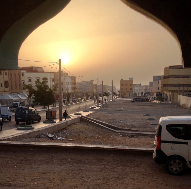 A dusty sunset over a busy and dirty looking street, which stretches into the distance. The photo is mostly composed of orange colours, and the domed silhouette of the hotel doorway obscures the top quarter of the image.