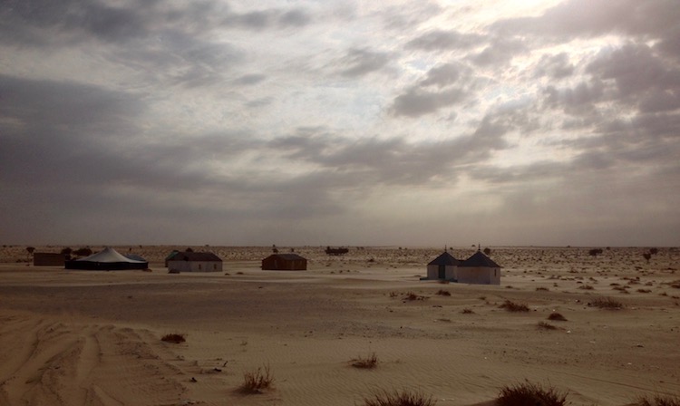 A dark, cloudy, brooding sky over various, sparsely distributed, small huts and tents in Mauritania. Surrounding them is desert. The sun is just breaking through some small gaps in the clouds, but the scene is still gloomy.