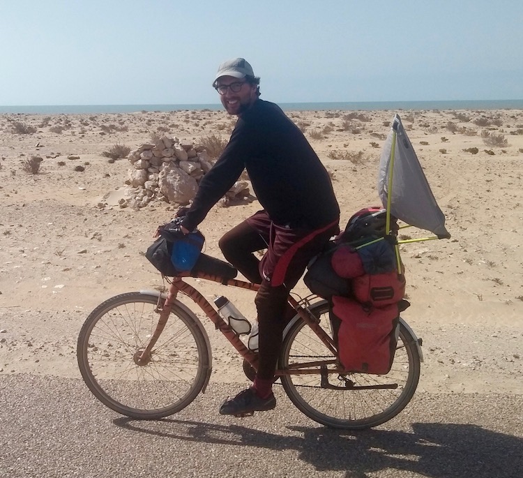 Side-on photo of Dan riding his bike, with a constructed helinox camping chair (chair zero) strapped to the luggage on the rear, acting as a wind sail.