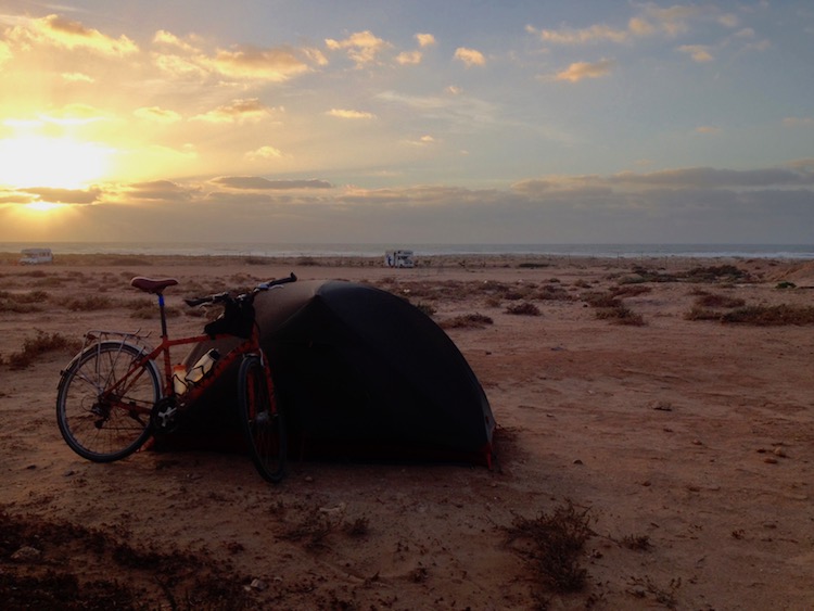 Dan's tent and bike parked on a flat beach, perhaps 150 meters from the sea. The sun is setting over the ocean and the sky is mostly clear, except for a long cloud in the distance.