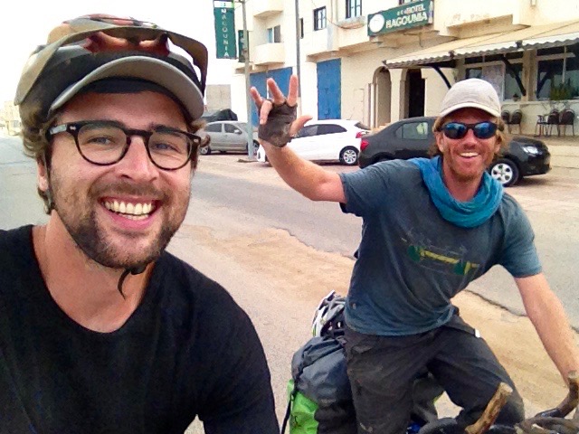 Dan and Emmanuel both riding their bikes and smiling at the camera (held by Dan). Dan is wearing a helmet and Emmanuel is wearing a cap and sunglasses.