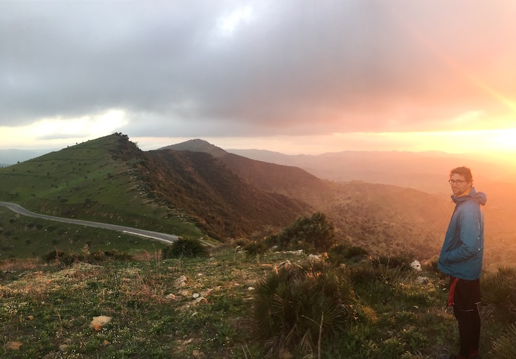 Mountaintop photo of Dan, looking at the camera, in front of a golden sunset. A small mountain road can also been seen weaving among the peaks.