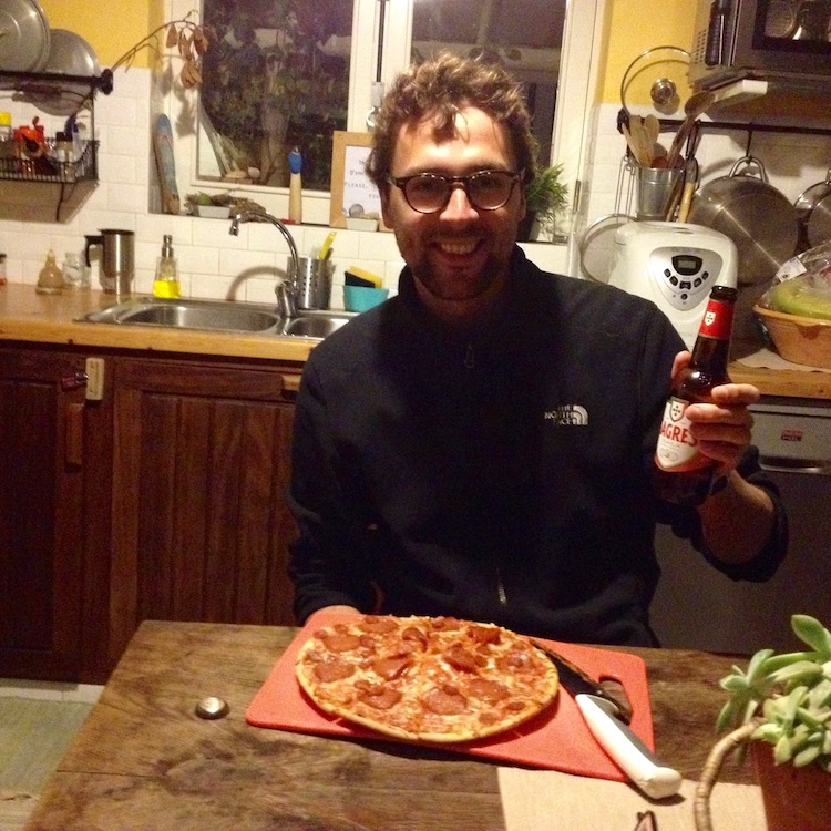 Dan sitting at a table in a cosy kitchen, happily holding up a Sagres beer, with a cooked pizza in front of him on a chopping board.