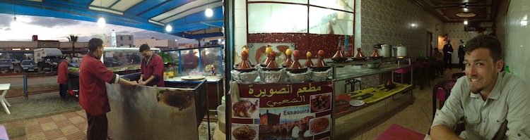 Wide panorama full of detail. On the left, the street can be seen, then the glowing bulds of the restaurant from which the photo is taken, then the chefs preparing food over grills, then an array of tagines sitting over fires, then the rear of the restaurant and then Nico sitting at the table with a hungry smile.