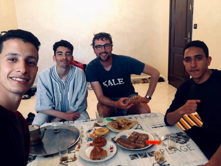 Dan with three Moroccan students, sitting on stools around a low table. They are smiling at the camera and on the table is an assortment of pastries (Dan is also holding one).