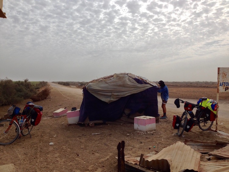 In between the dense thicket of reeds (from the banks of the river) and the road (which is a dirt track), there is a small makeshift hut, surrounding by Dan's, Flo's and Leo's touring bikes. Flo is standing next to the hut, trying to make it windproof.