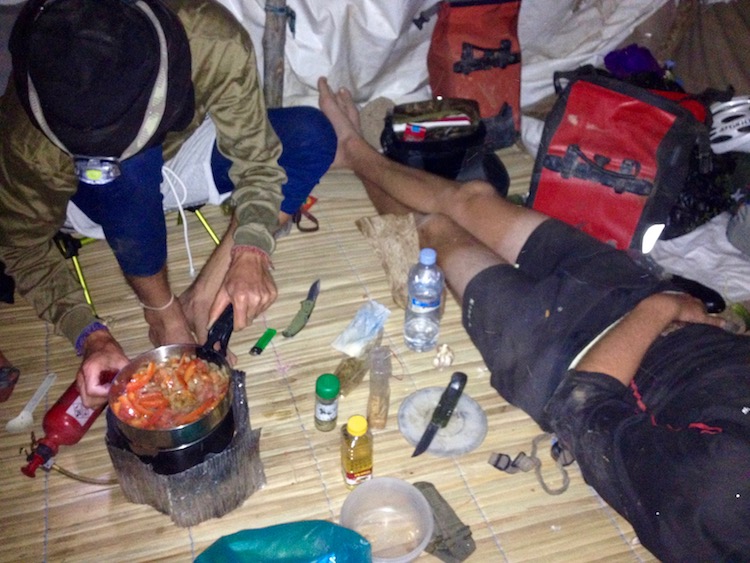 Flo bent over a stove, with a colourful mix of vegetables frying in a pan on the stove. Leo is reclined next to the stove. There is a bamboo mat on the floor. Photo taken from inside the makeshift hut.