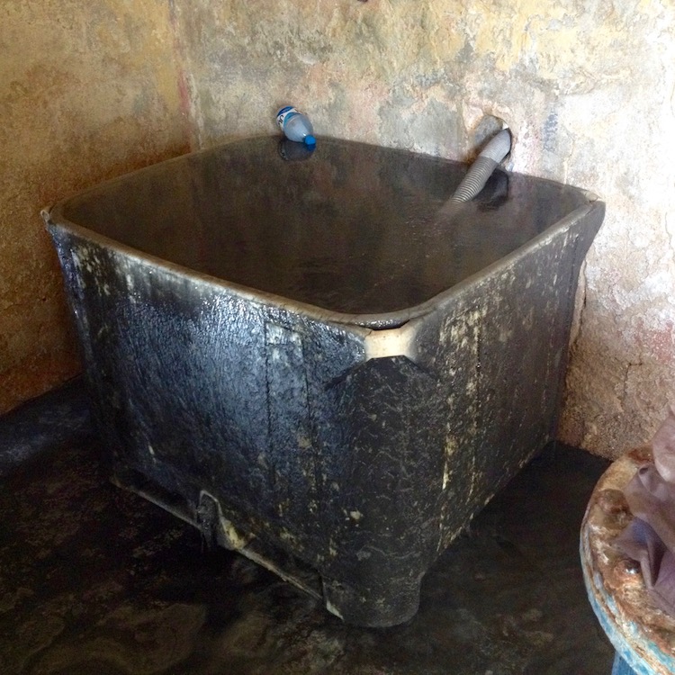 A large, sulphur-blackened tub, filled with murky, black water, in the corner of a concrete building. A pipe is running through one of the walls and into the tub.