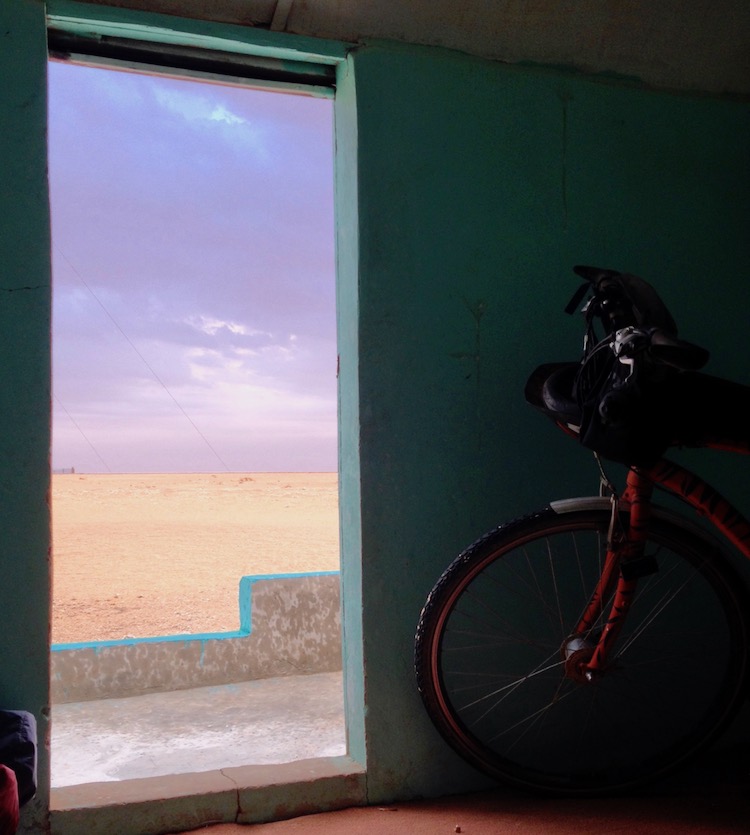 Photo taken from inside a hut, looking out of the open door. The sky is a dark purple colour, with clouds heavy with rain, and the landscape is sandy desert. The inside of the hut is painted light blue.
