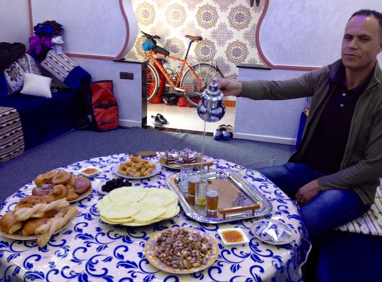 Ismain sitting at the table pouring tea and looking cool. He is holding the kettle about a foot above the tiny cup, with the tea streaming out directly into the cup, and he is also looking into the camera.