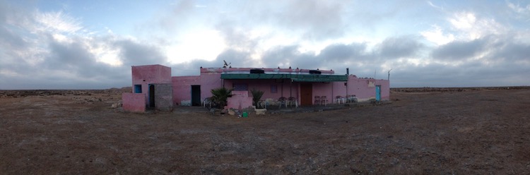 Panorama image of a one-storey building (painted light pink) surrounded by desert plain.