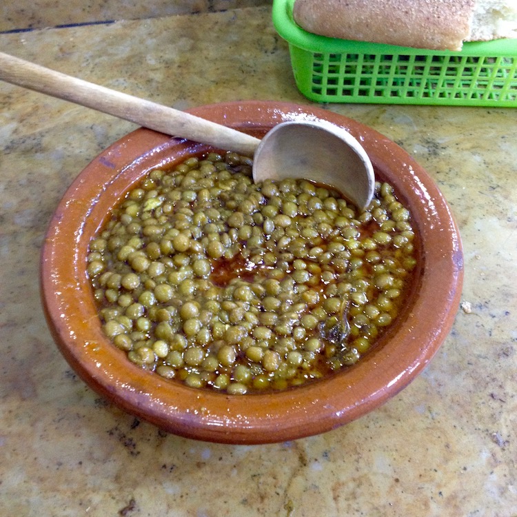Green lentils in an oily sauce, sitting in a brown tagine dish (moroccan traditional dish), with a wooden spoon sitting in the bowl too. Some bread can be seen in the background.