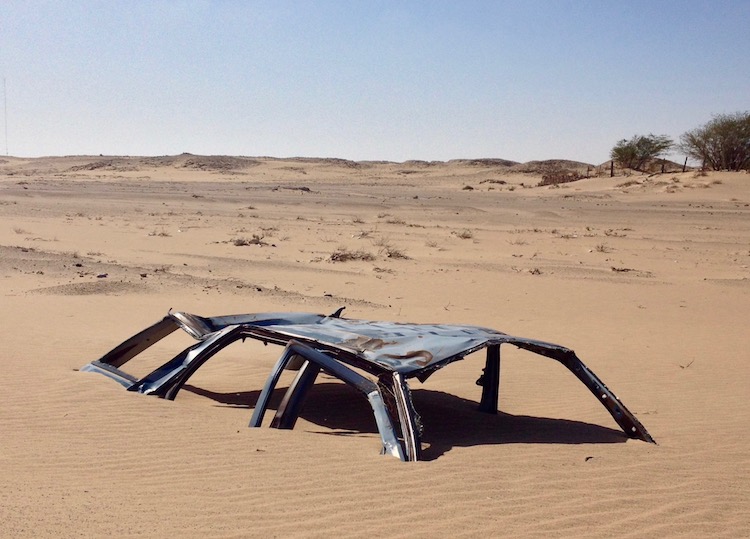 Only the roof and window struts of a car wreck can be seen above the sand, giving the impression of a rusty spider standing on the sand.