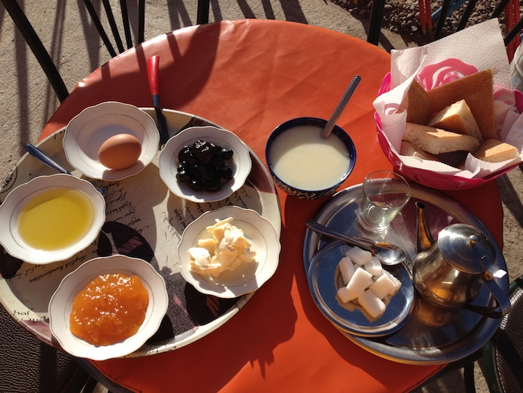 Assorted condiments in small porcelain dishes, on a silver tray. A black bowl filled with porridge, a silver tea set with sugar and a bowl with triangles of bread in it.
