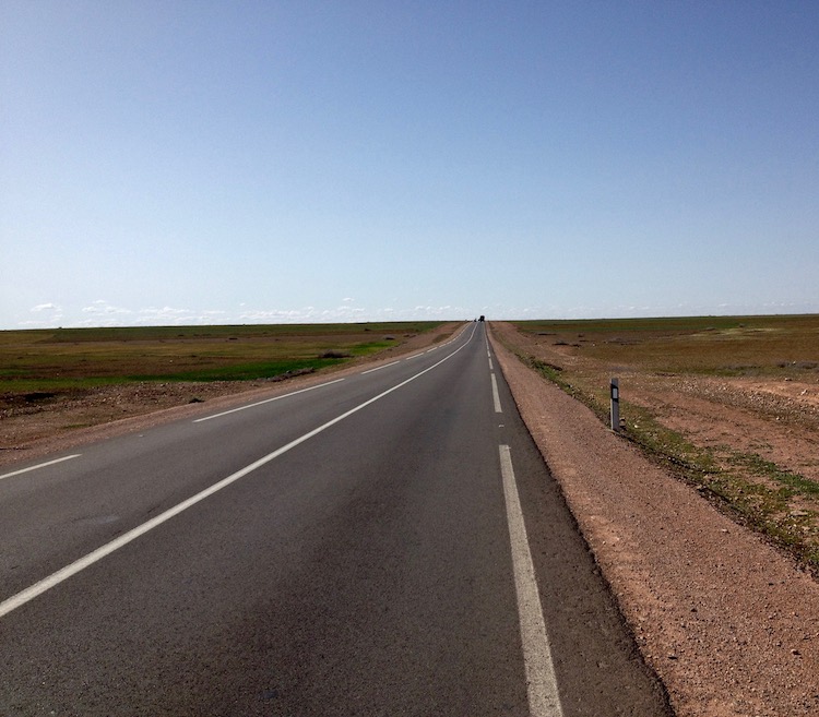 A simply, straight road with two lanes. Either side of the road, the ground is completely flat as far as the eye can see, with occasional patches of scrubby grass.