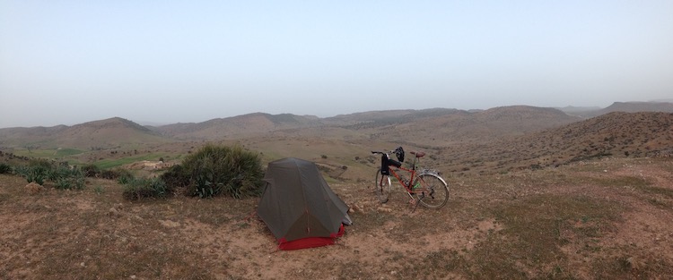 Dan's tent and bike on top of a hill, overlooking a sparse landscape of dusty sand and bushes (wild camping).