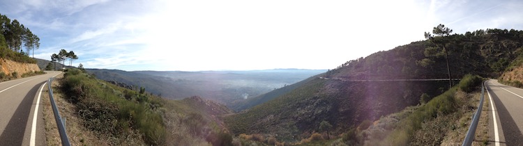A panorama showing a sunny road bending around the mountainside, with a view of the lower plain in the background