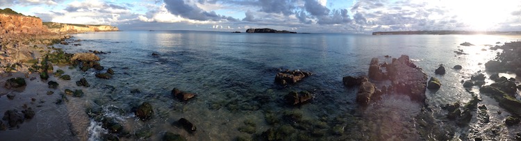 Panorama showing golden cliffs (lit by the sun) on one side, sun reflecting off the water on the other side and clear pools of turquoise water in the foreground, around the rocks.