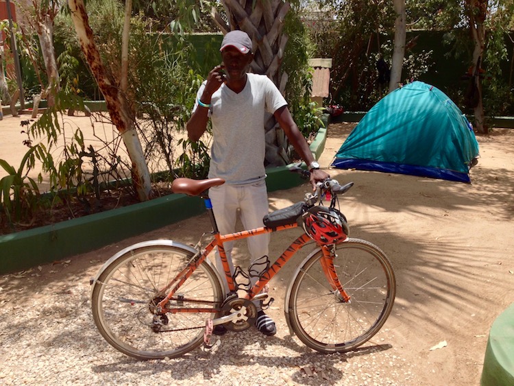 A Gambian local (Yusoupha), standing with my bike after purchasing it. In the background is a tent and the sandy ground of the campsite I was staying at.