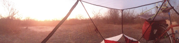 Panoramic photo from sitting inside my tent. Through the mesh, flat, dry, sandy ground can be seen, with dry bushes here and there. The sun has just risen and the colours are mostly orange.