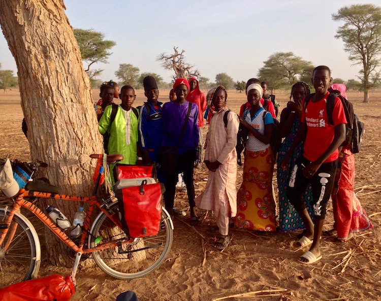 My bike leaning against a tree on a large, open, plain, with about fifteen school kids (dressed in colourful clothes) standing next to it, grinning at the camera. In the background are other trees, sparsely separated.