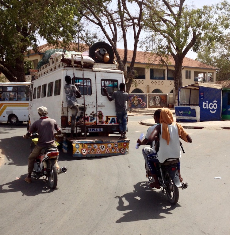 Photo taken while cycling around a roundabout in Thiès. Two motorbikes are in front, and in front of them is a white minivan (bus), with two young Senegalese people standing and hanging on to the back.