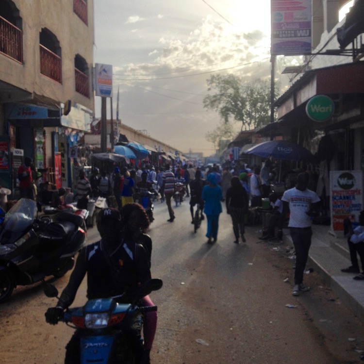 A small, but very busy and bustling street, with many shops and stalls on either side. It is late afternoon, and the sun is breaking through some gaps in the clouds, to give the scene quite an atmospheric look.