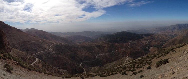 Panorama looking down on a huge view, mountains descend in the foreground and a plain stretches out to the horizon. A road can be seen snaking like spaghetti across all the mountains.