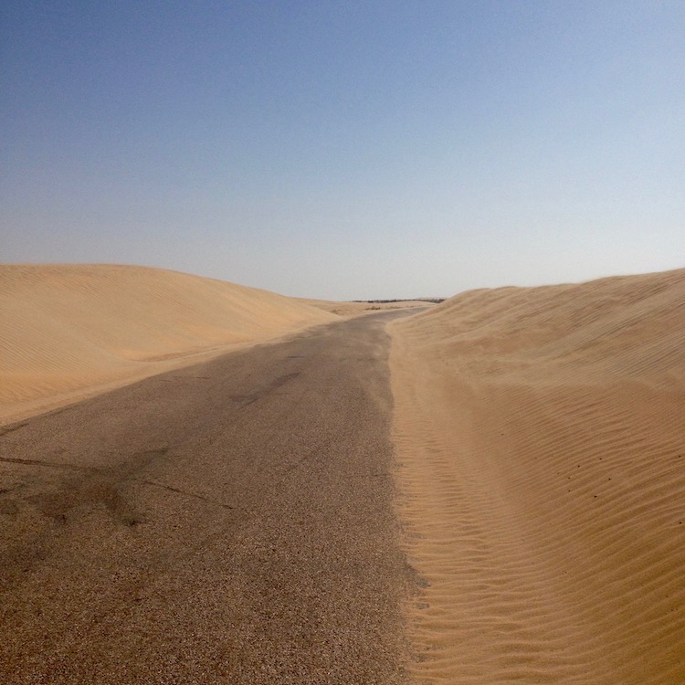 A plain road, with no markings, running into the distance, with a small sand dune (~2m) piled up on either side.