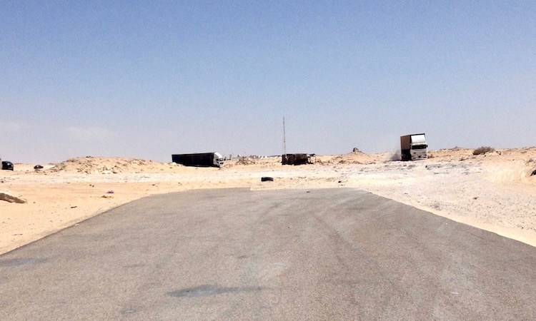 Photo taken from a road, showing the road ending abruptly and turning into a rocky, sandy piste. In the distance, there are a few lorries turned side-on, searching for a path.