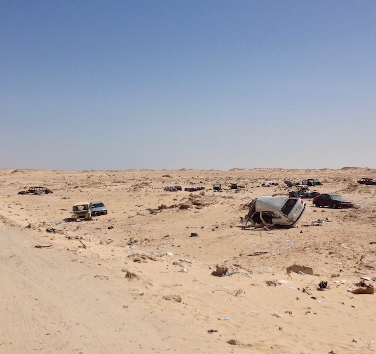 Sandy wasteland, with various car wrecks, some more intact than others. The ground is also littered with smaller car parts.