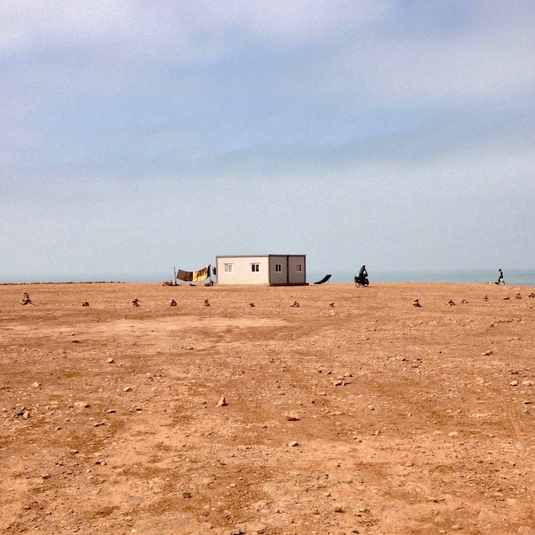 Flat sandy ground, stretching out towards a cliff edge (the sea can be seen beyond that) with a small, cubic, prefabricated bungalow sitting about 30 meters from the edge. Clothes can be seen drying on one side, and the silhouette of Emmanuel riding his bike can be seen on the other (this photo is taken from a distance).