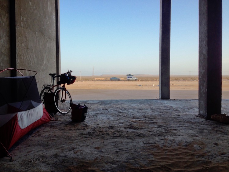 Looking out from the back of a large, unfinished room of a gas station. The ceiling is so high that it is not seen in the photo. Dan's tent is half set up against the wall on the left, and in the background is a road running by, with some cars on it, then the desert plain, stretching to the horizon.