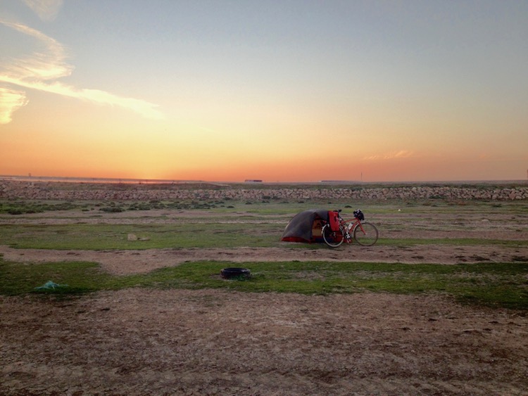 Dan's tent with bike propped up next to it, in a dry river bed. The mud is cracked and there are some patches of short grass. The sun has just set, making the sky a pink colour.